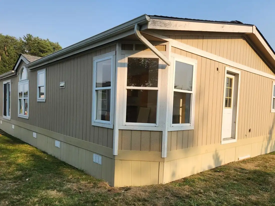 Manufactured home freshly painted tan with white trim, a white bay window and two-tone skirting