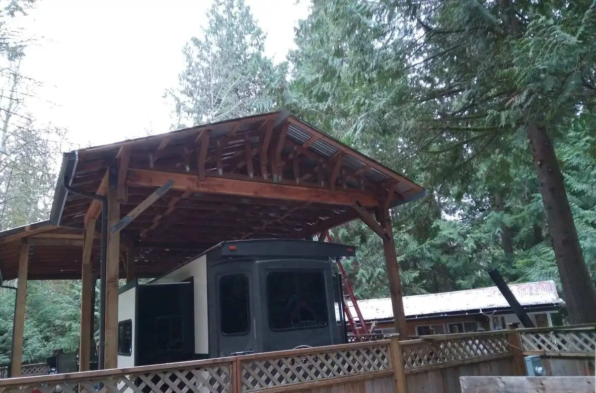 Timber-frame roof on knee-braced posts with exposed trusses, sheltering a travel trailer and its deck among tall evergreens, Sedro-Woolley